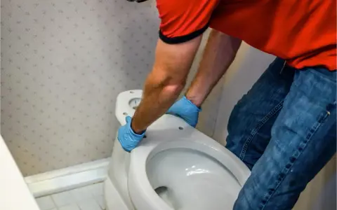 Hombre con guantes azules desmonta asiento de inodoro. Aprende como quitar un bidet para renovar tu baño.
