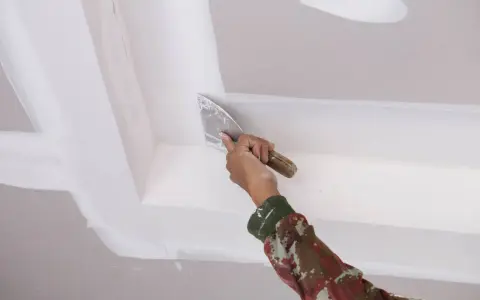 Mano con espátula aplica masilla para reparar techo escayola, dejando un acabado liso y perfecto.