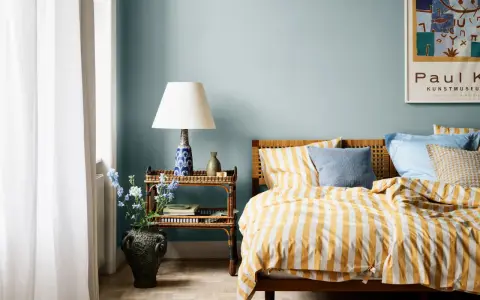 Habitación con paredes azul claro, cama con ropa de cama a rayas amarillas y blancas, y mesita de noche de ratán. Ideas de colores para habitaciones.
