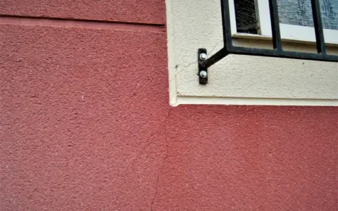 Grietas profundas en pared de color rojo, cerca de una ventana con rejas. Aprende como arreglar grietas profundas en paredes.