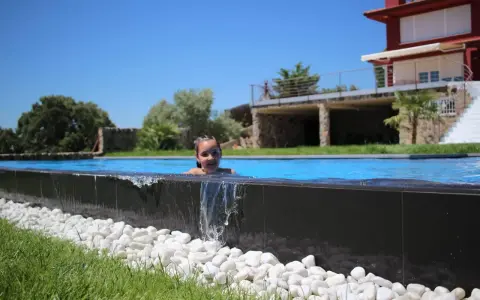Niña sonriente en una piscina desbordante, con agua cayendo sobre piedras blancas y césped verde. Casa moderna al fondo.
