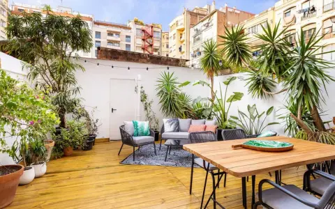 Un patio exterior pequeño transformado con una reforma: suelo de madera, muebles de exterior, plantas exuberantes y luces decorativas crean un oasis urbano.