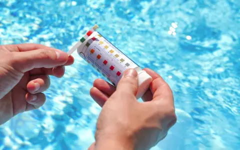 Manos sostienen una tira reactiva para medir el pH de la piscina. El agua azul y brillante de la piscina se ve al fondo.