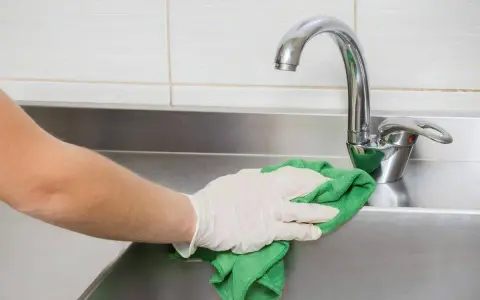 Mano con guante limpiando fregadero de acero inoxidable con paño verde.