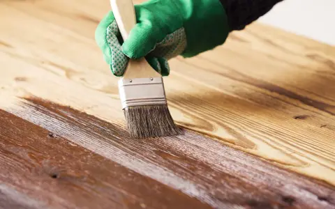 Mano con guante verde aplicando barniz a madera. El proceso de barnizar madera protege y embellece la superficie.