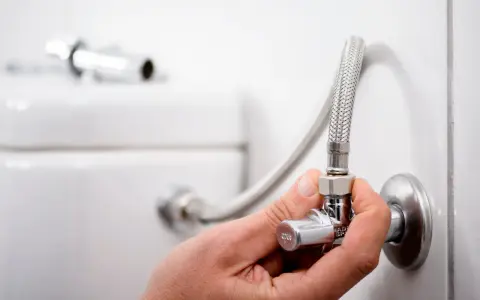 Mano ajustando uno de los tipos de llaves de paso de agua, conectada a una manguera flexible y al tanque de un inodoro.