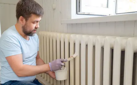 Hombre con barba pintando radiadores blancos con brocha y pintura.