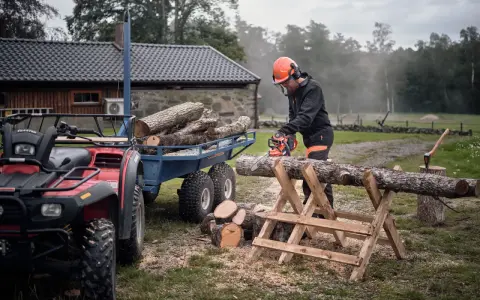 Hombre corta leña con motosierra, pensando cuál es la mejor leña para chimenea. Remolque con troncos y ATV al lado.