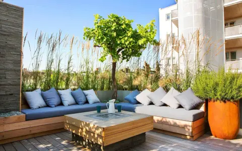 Elegante decoracion terrazas aticos con sofás de madera, cojines grises y un centro de mesa con fuego. Hierbas altas y un árbol crean un oasis urbano.