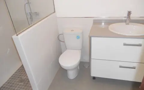 Baño moderno con ducha separada por muro de azulejos blancos y cristal. Inodoro y lavabo sobre mueble blanco.