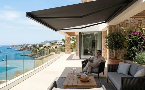 Hombre relajado en terraza con toldo oscuro, disfrutando de la vista al mar. Los colores de toldos de moda crean un ambiente moderno y elegante.