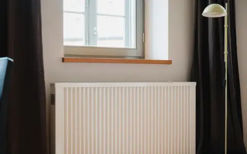 Radiador blanco bajo ventana, con cortinas oscuras y lámpara amarilla. El **calor azul** del invierno se combate con este sistema.