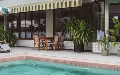 Terraza con piscina y toldo a rayas verdes y blancas. Mesa de madera con sillas de mimbre listas para instalar toldo.
