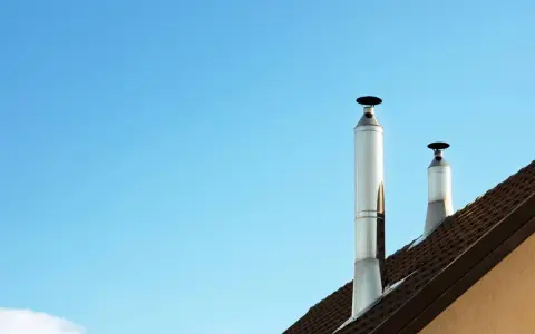 Dos tubos de chimenea metálicos se alzan sobre un tejado marrón contra un cielo azul claro. Es un día perfecto para aprovechar el calor del tubo de chimenea.