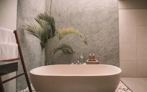 Un baño moderno con una bañera ovalada, toallas blancas en una escalera de madera y una planta de hojas verdes. La pared tiene textura y el suelo está cubierto de guijarros. ¡Listo para pintar la bañera y renovar el espacio!