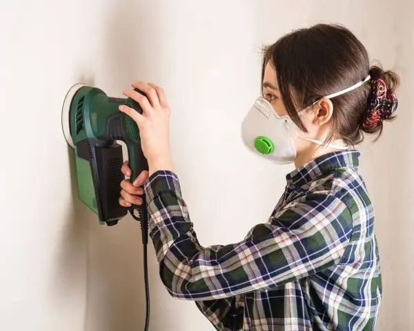 Mujer con mascarilla lijando paredes con una lijadora eléctrica.