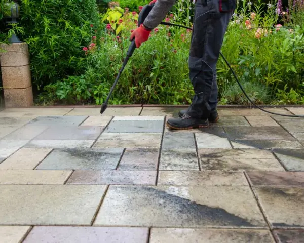 Persona con guantes rojos usa hidrolimpiadora para limpiar suelo de terraza exterior con baldosas de piedra.