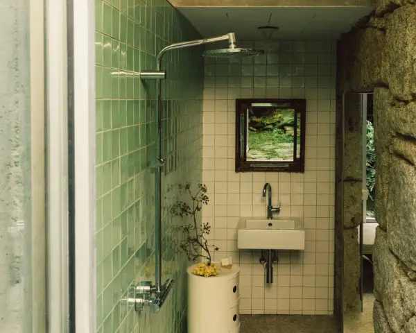 Baños pequeños con ducha, lavabo y espejo con vista a un jardín rocoso. Paredes de azulejos verdes y piedra natural.