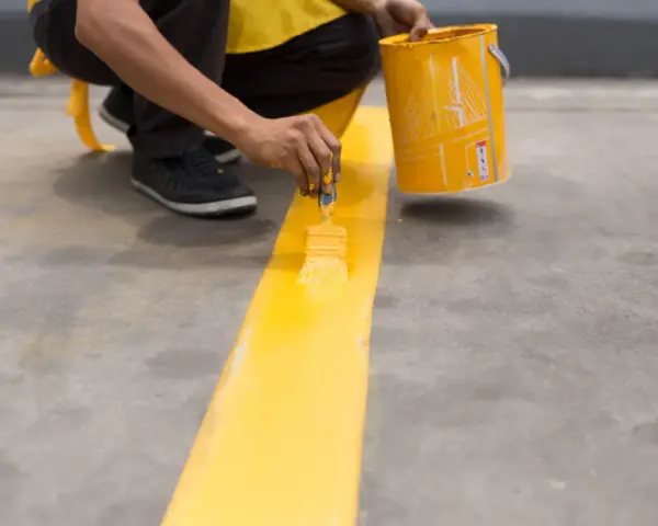 Persona pintando una línea amarilla sobre suelo de cemento sin pulir, con una lata de pintura al lado.