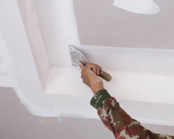 Mano con espátula aplica masilla para reparar techo escayola, dejando un acabado liso y perfecto.
