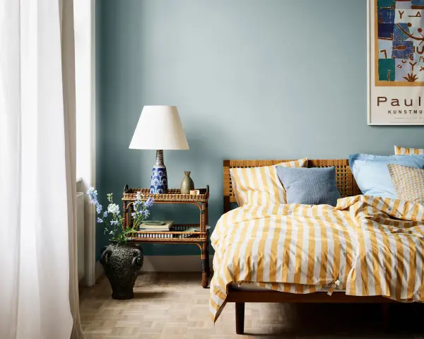 Habitación con paredes azul claro, cama con ropa de cama a rayas amarillas y blancas, y mesita de noche de ratán. Ideas de colores para habitaciones.