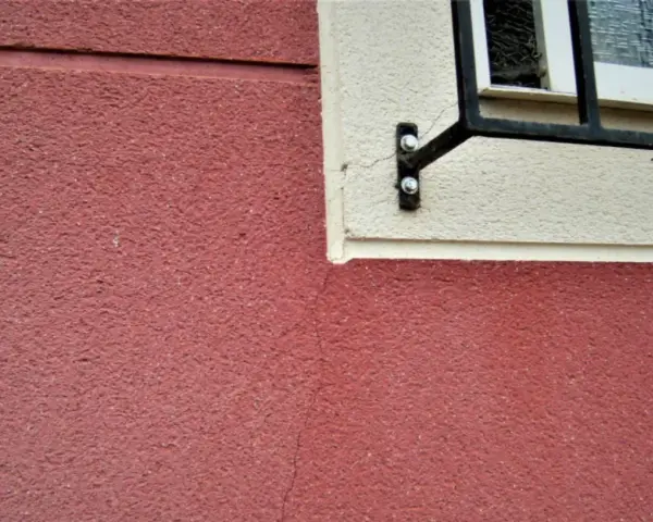 Grietas profundas en pared de color rojo, cerca de una ventana con rejas. Aprende como arreglar grietas profundas en paredes.