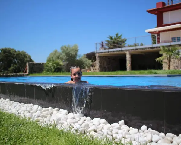 Niña sonriente en una piscina desbordante, con agua cayendo sobre piedras blancas y césped verde. Casa moderna al fondo.