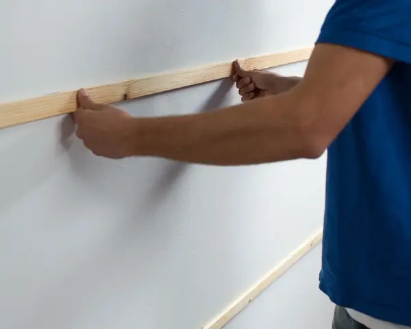 Man placing wooden slats on a white wall, demonstrating como poner listones de madera en la pared.