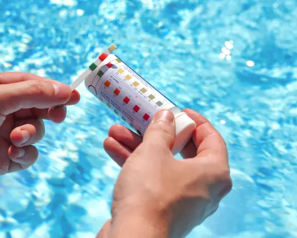 Manos sostienen una tira reactiva para medir el pH de la piscina. El agua azul y brillante de la piscina se ve al fondo.