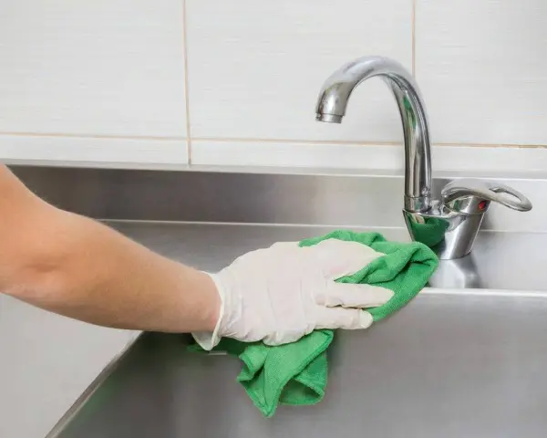 Mano con guante limpiando fregadero de acero inoxidable con paño verde.