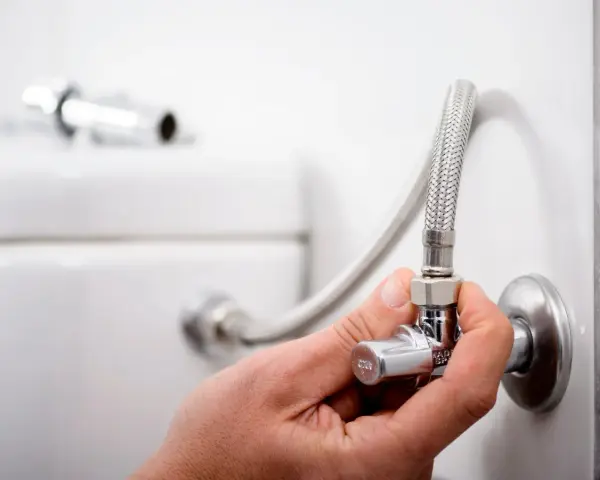 Mano ajustando uno de los tipos de llaves de paso de agua, conectada a una manguera flexible y al tanque de un inodoro.