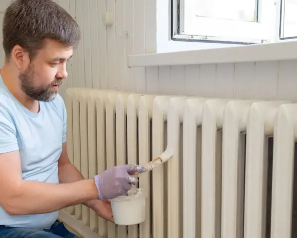 Hombre con barba pintando radiadores blancos con brocha y pintura.