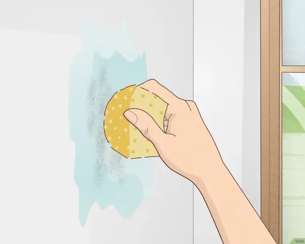 Mano limpiando una mancha en pared blanca con esponja y agua. Aprende como limpiar paredes blancas fácilmente.