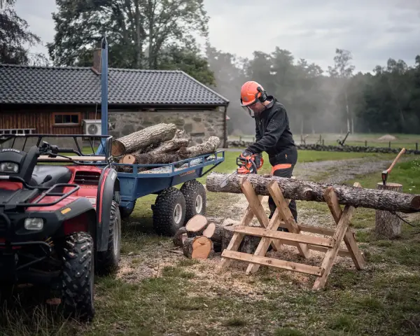 Hombre corta leña con motosierra, pensando cuál es la mejor leña para chimenea. Remolque con troncos y ATV al lado.