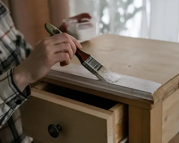 Persona con camisa a cuadros pinta madera de un mueble con brocha blanca.
