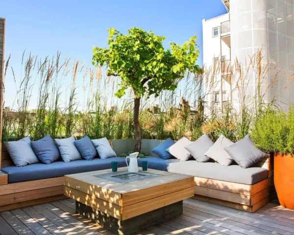 Elegante decoracion terrazas aticos con sofás de madera, cojines grises y un centro de mesa con fuego. Hierbas altas y un árbol crean un oasis urbano.