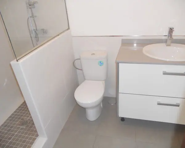 Baño moderno con ducha separada por muro de azulejos blancos y cristal. Inodoro y lavabo sobre mueble blanco.