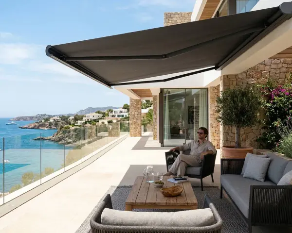 Hombre relajado en terraza con toldo oscuro, disfrutando de la vista al mar. Los colores de toldos de moda crean un ambiente moderno y elegante.