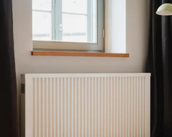 Radiador blanco bajo ventana, con cortinas oscuras y lámpara amarilla. El **calor azul** del invierno se combate con este sistema.