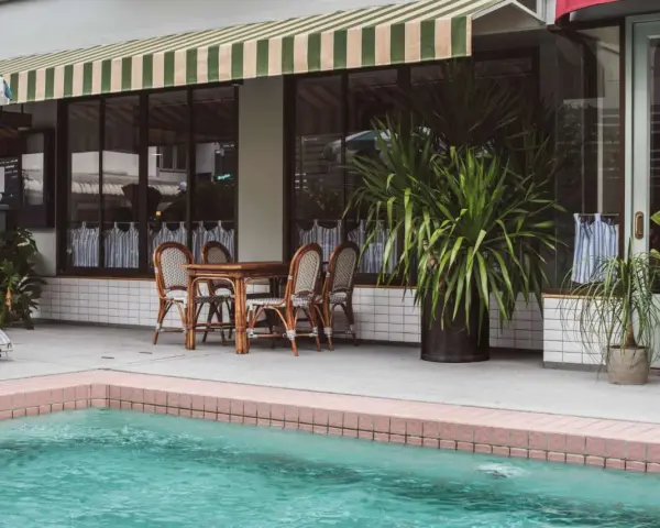 Terraza con piscina y toldo a rayas verdes y blancas. Mesa de madera con sillas de mimbre listas para instalar toldo.