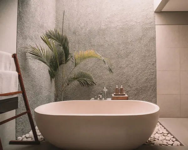Un baño moderno con una bañera ovalada, toallas blancas en una escalera de madera y una planta de hojas verdes. La pared tiene textura y el suelo está cubierto de guijarros. ¡Listo para pintar la bañera y renovar el espacio!