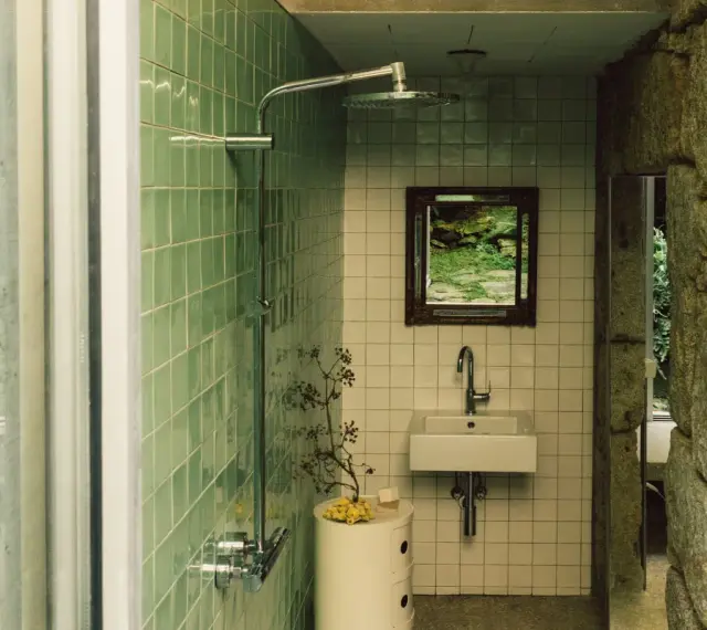 Baños pequeños con ducha, lavabo y espejo con vista a un jardín rocoso. Paredes de azulejos verdes y piedra natural.