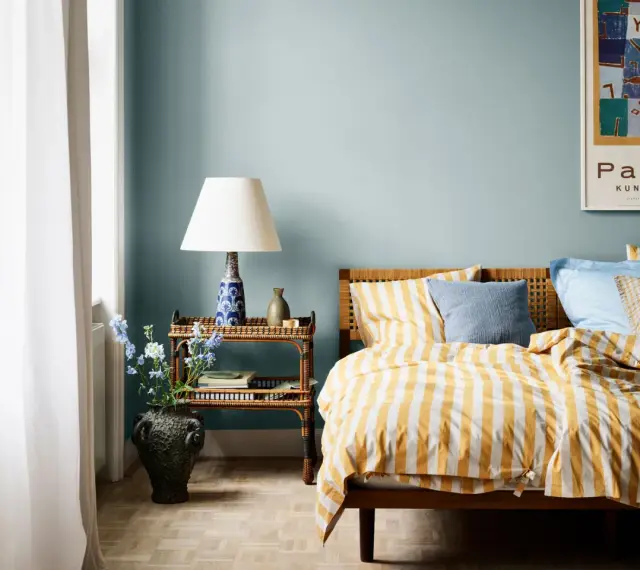 Habitación con paredes azul claro, cama con ropa de cama a rayas amarillas y blancas, y mesita de noche de ratán. Ideas de colores para habitaciones.