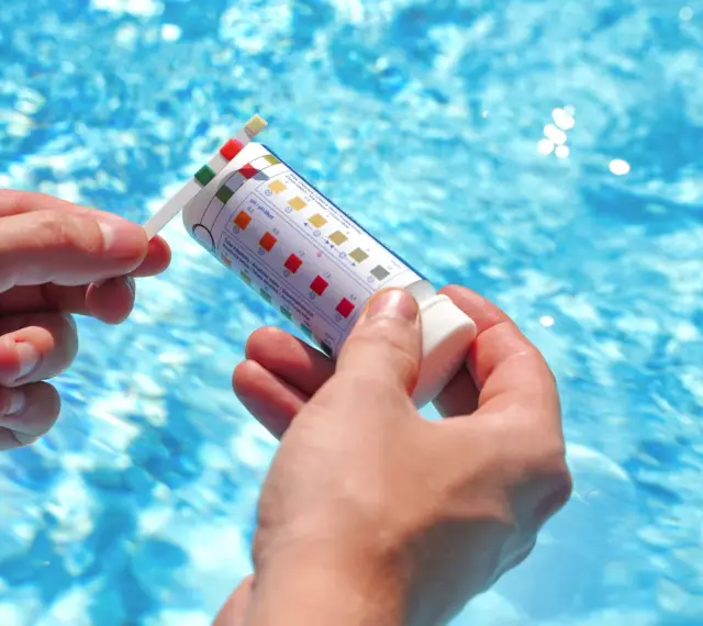 Manos sostienen una tira reactiva para medir el pH de la piscina. El agua azul y brillante de la piscina se ve al fondo.