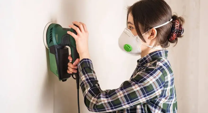 Mujer con mascarilla lijando paredes con una lijadora eléctrica.