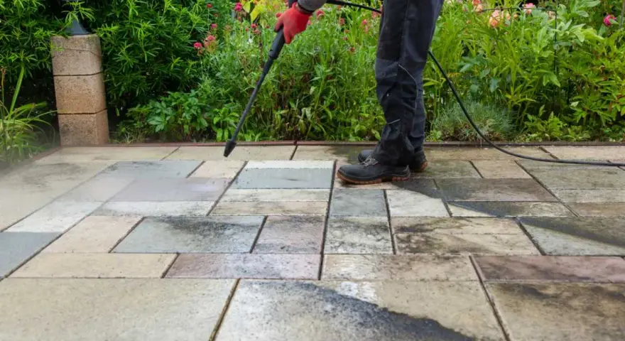Persona con guantes rojos usa hidrolimpiadora para limpiar suelo de terraza exterior con baldosas de piedra.