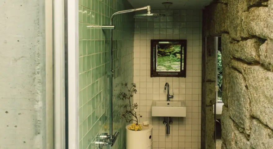 Baños pequeños con ducha, lavabo y espejo con vista a un jardín rocoso. Paredes de azulejos verdes y piedra natural.