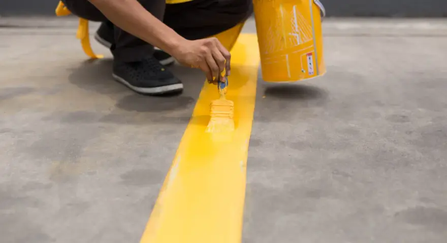 Persona pintando una línea amarilla sobre suelo de cemento sin pulir, con una lata de pintura al lado.