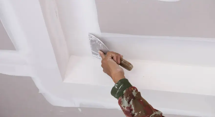 Mano con espátula aplica masilla para reparar techo escayola, dejando un acabado liso y perfecto.