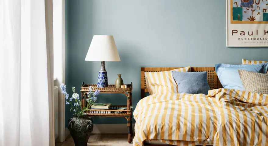Habitación con paredes azul claro, cama con ropa de cama a rayas amarillas y blancas, y mesita de noche de ratán. Ideas de colores para habitaciones.